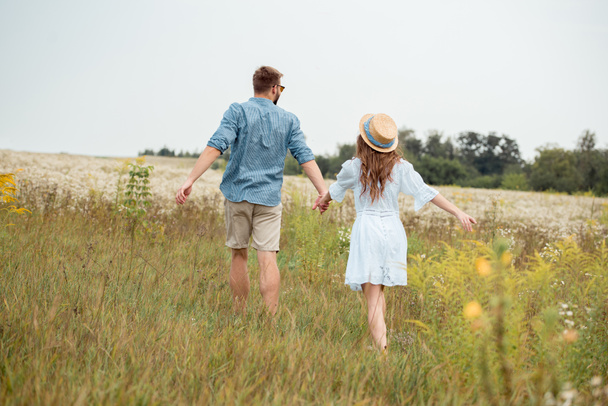 back view of young lovers holding hands while running in field with wild flowers - Photo, image