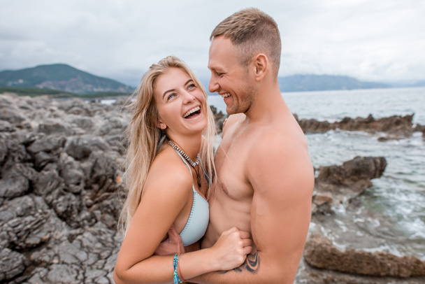 вид збоку щасливі молода пара, обіймати і сміючись кам'янистий пляж у Чорногорії - Фото, зображення