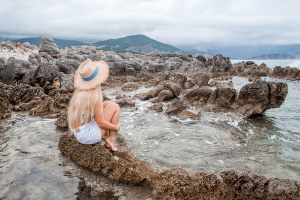 κορίτσι στο μπικίνι και ψάθινο καπέλο που κάθεται στην βραχώδη παραλία του Μαυροβουνίου - Φωτογραφία, εικόνα