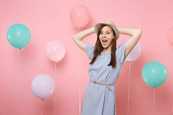 Portret van dolblij gelukkig jonge vrouw dragen van stro zomer hoed en blauwe jurk houden handen in de buurt van hoofd op roze achtergrond met kleurrijke lucht ballonnen. Vakantie verjaardagspartij mensen oprechte emoties - Foto, afbeelding