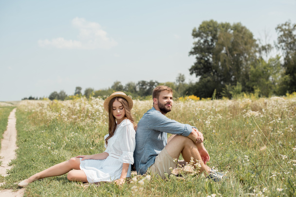 pareja joven descansando espalda con espalda en la manta en el campo con flores silvestres en el día de verano
 - Foto, Imagen