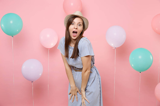 Portrait of shocked amazed young woman wearing straw summer hat and blue dress standing on pastel pink background with colorful air balloons. Birthday holiday party people sincere emotions concept - Foto, Imagem