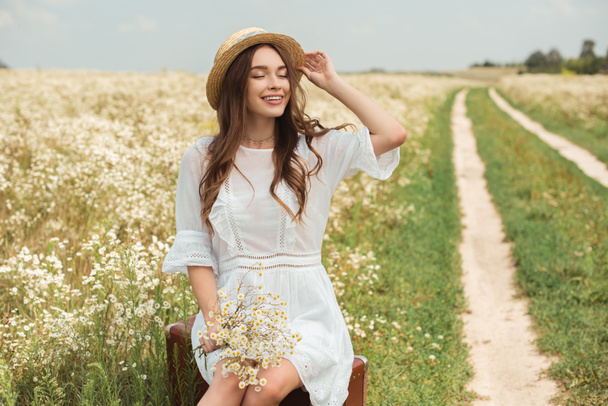 mujer sonriente en vestido blanco con ramo de flores de manzanilla silvestre sentado en la maleta retro en el campo
 - Foto, Imagen