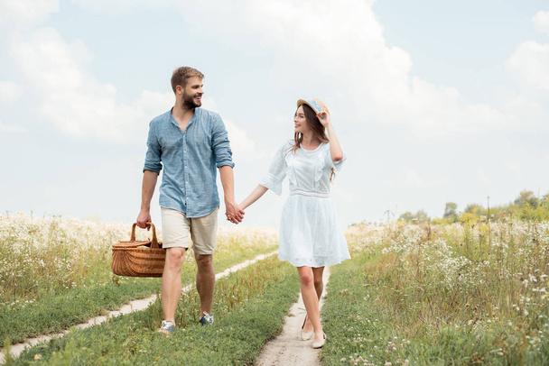 sonriente pareja con cesta de picnic cogido de la mano mientras camina en el campo
 - Foto, Imagen