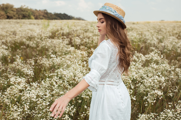 pretty pensive woman in straw hat and white dress on meadow with wild flowers - 写真・画像