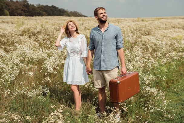 happy couple with retro suitcase holding hands while standing in field with wild flowers - Fotó, kép
