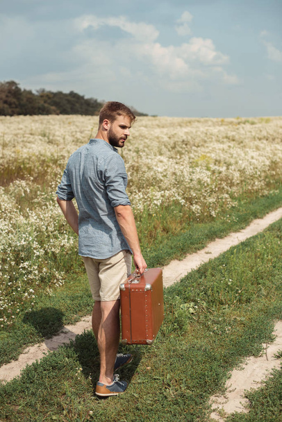 vista posterior del hombre con maleta retro de pie en archivado con flores silvestres en el día de verano
 - Foto, Imagen