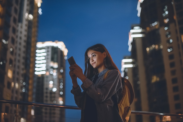 улыбающаяся женщина с рюкзаком с помощью смартфона на улице с ночным городским освещением на заднем плане
 - Фото, изображение