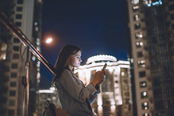 боковой вид привлекательной женщины с помощью смартфона на улице с ночными городскими огнями на заднем плане
 - Фото, изображение