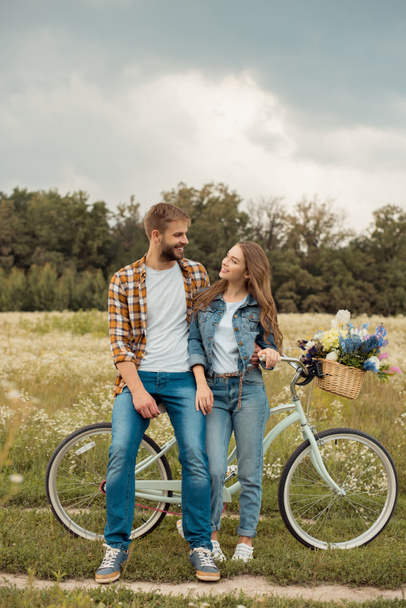 amantes sorridentes com bicicleta retro olhando um para o outro no campo com flores selvagens
 - Foto, Imagem