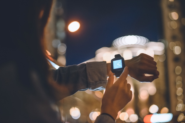 vista parcial de la mujer con smartwatch en la muñeca y luces de la ciudad nocturna en el fondo
 - Foto, imagen
