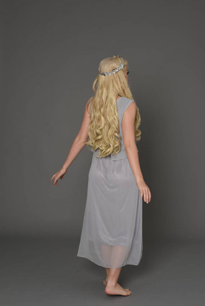 full length portrait of blonde girl wearing dress and crown. standing pose with back to the camera. grey studio background. - Foto, Bild