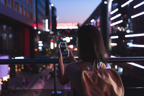 rear view of woman with backpack and smartphone with booking website on screen in hands standing on night city street - 写真・画像