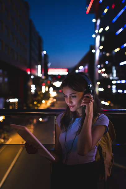 портрет женщины в наушниках с помощью планшета на улице с ночным городским освещением на заднем плане
 - Фото, изображение
