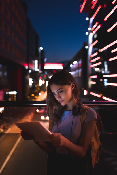 portrait of young woman with headphones using tablet on street with night city lights on background - Φωτογραφία, εικόνα