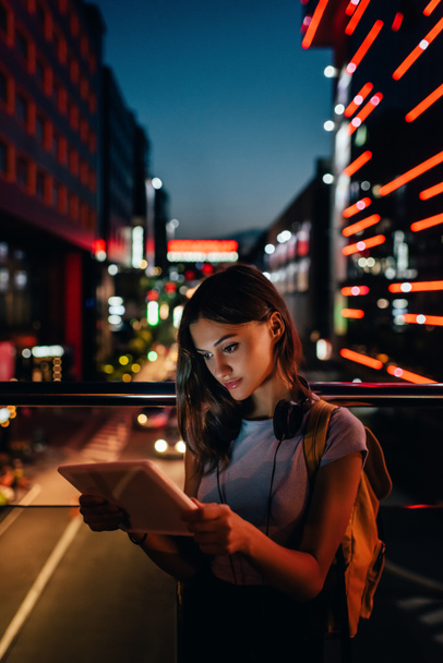 портрет молодой женщины в наушниках с помощью планшета на улице с ночным городским освещением на заднем плане
 - Фото, изображение