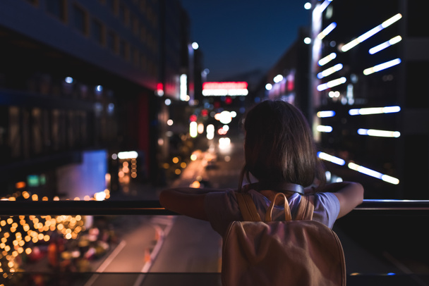 Вид сзади женщины с рюкзаком, стоящей на улице и смотрящей на ночные городские огни
 - Фото, изображение