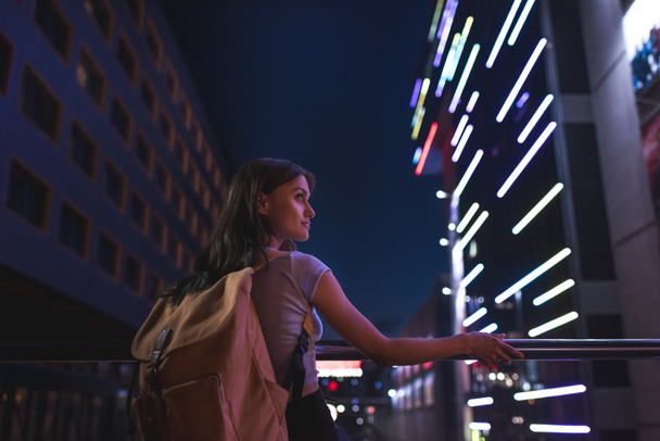 красивая застенчивая женщина с рюкзаком, смотрящая вдаль по ночной улице города
 - Фото, изображение