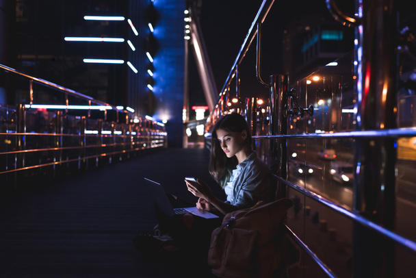 Seitenansicht einer jungen Frau mit Laptop auf Knien und Smartphone mit nächtlicher Stadt im Hintergrund - Foto, Bild