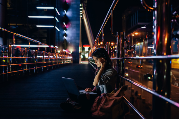 Μερική άποψη του γυναίκα ακούγοντας μουσική στα ακουστικά ενώ χρησιμοποιείτε φορητό υπολογιστή στο δρόμο τη νύχτα της πόλης - Φωτογραφία, εικόνα