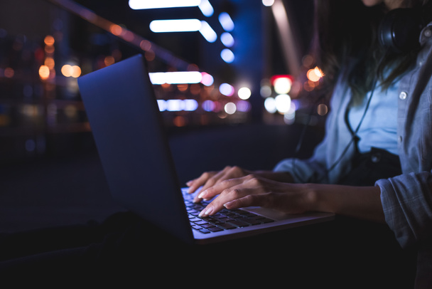 recortado disparo de mujer con auriculares escribiendo en el ordenador portátil en la calle de la ciudad por la noche
 - Foto, Imagen