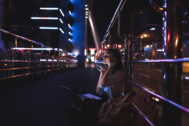 Widok z boku kobieta słuchania muzyki w słuchawkach z laptopem na kolanach na ulicy miasta w nocy - Zdjęcie, obraz