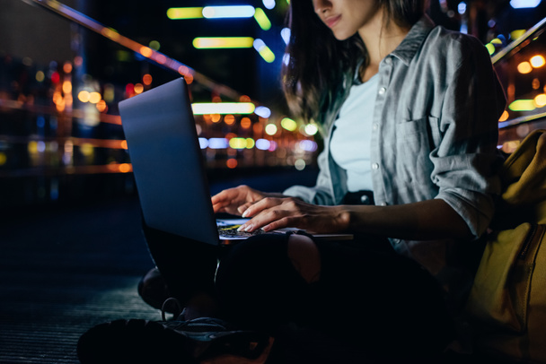 обрізаний знімок жінки, використовуючи ноутбук на вулиці з нічними вогнями міста на фоні
 - Фото, зображення