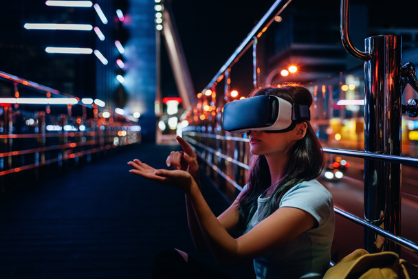 νεαρή γυναίκα στο σετ κεφαλής εικονικής πραγματικότητας που κάθεται στο δρόμο με την πόλη το βράδυ σε φόντο - Φωτογραφία, εικόνα