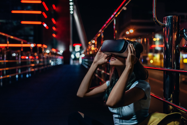 junge Frau im Virtual-Reality-Headset auf der Straße sitzend mit nächtlicher Stadt im Hintergrund - Foto, Bild