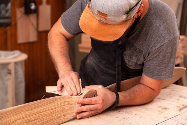 Erfahrener Tischler in Arbeitskleidung und Kleinunternehmer, der in der Holzwerkstatt arbeitet, verwendet Schleifpapier zum Polieren der Helikopterblätter von Kindern am Arbeitstisch in der Werkstatt   - Foto, Bild