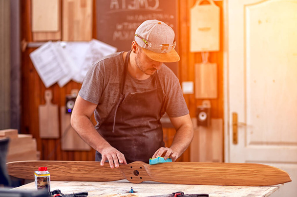 Ένας άνθρωπος της εργασίας σε ένα καπάκι και πουκάμισο γυαλίζει το ξύλινο μπλοκ με γυαλόχαρτο πριν από το βάψιμο στο εργαστήριο, στο παρασκήνιο, εργαλεία και μηχανήματα διάτρησης.  - Φωτογραφία, εικόνα