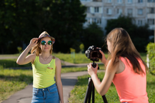 Two girlfriends schoolgirls in summer in the park records video on the camera. She is dressed in bright clothes with a hat and sunglasses. The concept of young bloggers. - Photo, image