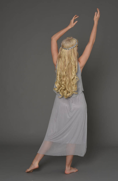 full length portrait of blonde girl wearing dress and crown. standing pose with back to the camera. grey studio background. - Photo, Image