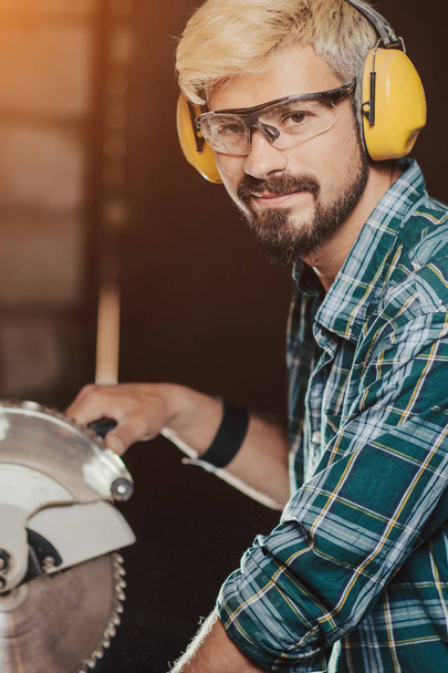 Bright young hipster bearded man with ear protectors by profession carpenter builder saws with a circular saw a wooden board on a wooden table in the workshop. - Foto, imagen