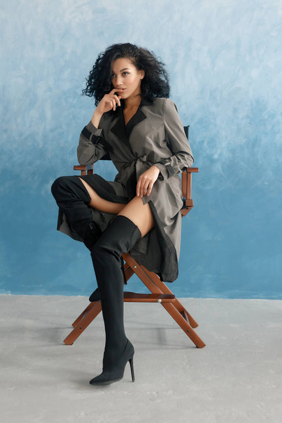 people, race, ethnicity and fashion concept - beauty black model posing in high boots. Young pretty beautiful mulatto woman sitting in director's chair at film. woman in gray raincoat, afro hairstyle - Zdjęcie, obraz