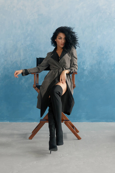 people, race, ethnicity and fashion concept - beauty black model posing in high boots. Young pretty beautiful mulatto woman sitting in director's chair at film. woman in gray raincoat, afro hairstyle - Valokuva, kuva
