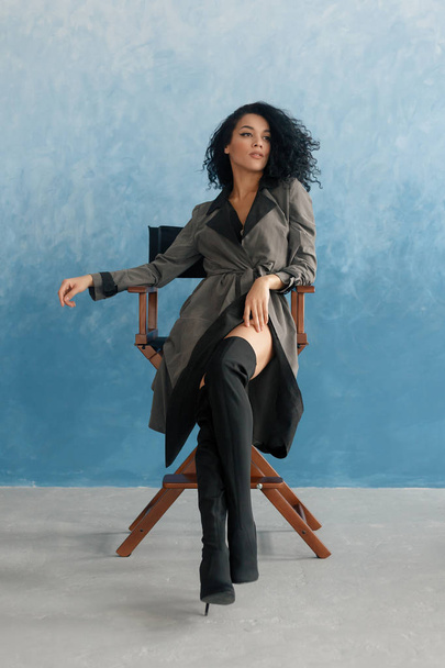 people, race, ethnicity and fashion concept - beauty black model posing in high boots. Young pretty beautiful mulatto woman sitting in director's chair at film. woman in gray raincoat, afro hairstyle - Photo, Image