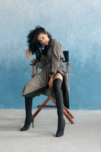 people, race, ethnicity and fashion concept - beauty black model posing in high boots. Young pretty beautiful mulatto woman sitting in director's chair at film. woman in gray raincoat, afro hairstyle - Фото, изображение