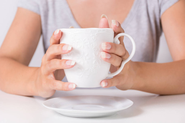 Young woman holding in hand white cup of coffee at home desk with natural manicure on finger nails. Beauty and drink concept. Close up, selective focus - Foto, immagini