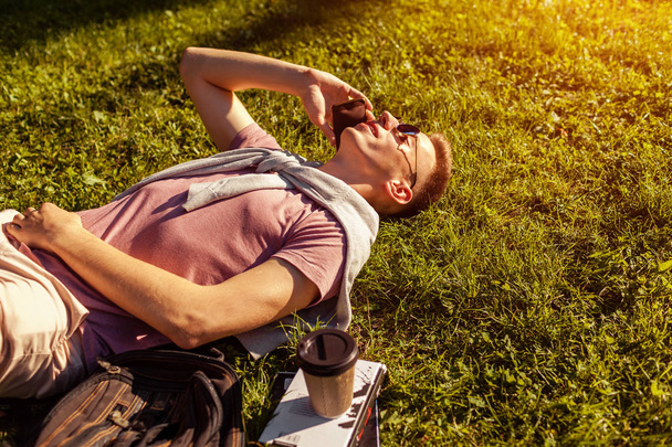 Handsome college man chilling in spring campus park. Happy guy student lying on grass talking on smartphone with book and coffee beside - Photo, image