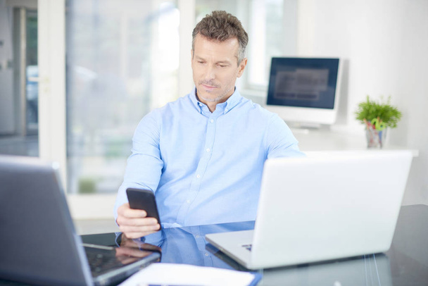 事務所で座っていると、作業中のシャツを着て思考のビジネスマンの肖像画。彼の携帯とノート パソコンでの作業中にメッセージ本文を使用してプロの男. - 写真・画像