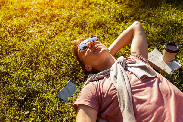 Красивий студент коледжу охолоджує у весняному кампусі парку. Щасливий хлопець студент лежить на траві зі своїм телефоном, книгою та кавою поруч під час заходу сонця
 - Фото, зображення