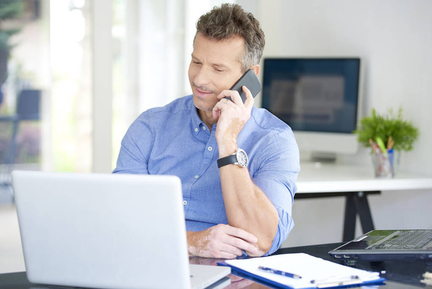 Portrait of middle aged businessman wearing shirt while sitting at the office and working. Professional man using his mobile phone and talking with somebody while working on laptops. Multitasking. - 写真・画像
