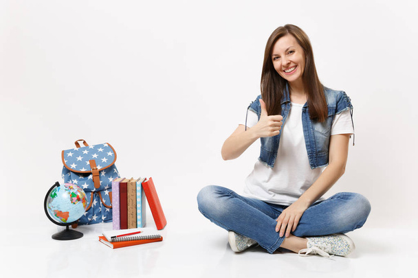 Портрет молодої випадкової усміхненої жінки-студентки в джинсовому одязі, що показує великий палець, що сидить поблизу глобуса, рюкзака, шкільних книг, ізольованих на білому тлі. Освіта в середній школі університетський коледж
 - Фото, зображення