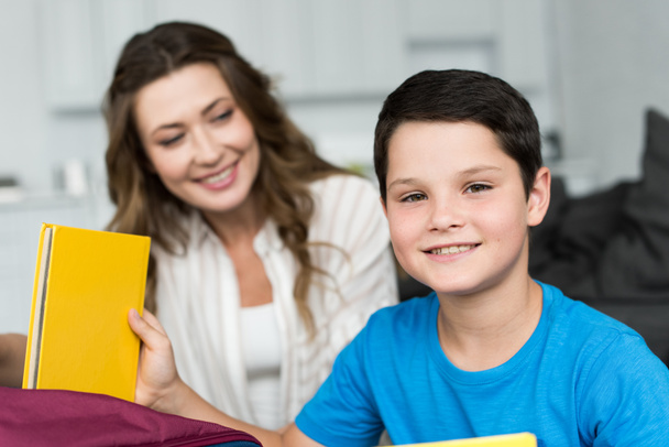 selective focus of smiling boy with book and mother behind at home - Fotografie, Obrázek