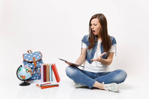 Шокована роздратована жінка-студент тримає за допомогою планшетного комп'ютера, розтягуючи руку, сидячи поблизу глобуса, рюкзака, шкільних книг ізольованих на білому тлі. Освіта в середній школі університетський коледж
 - Фото, зображення