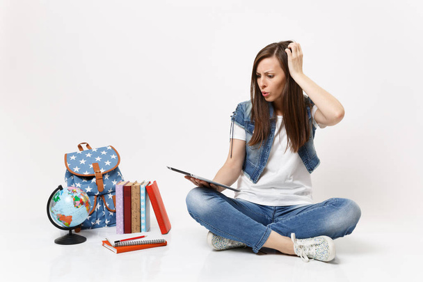 Concerned puzzled woman student holding using tablet pc computer, clinging to head, sitting near globe backpack, school books isolated on white background. Education in high school university college - Φωτογραφία, εικόνα