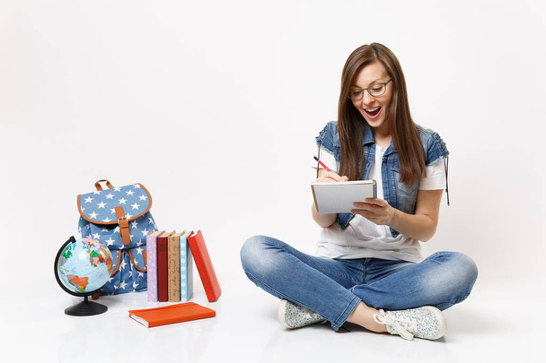 Молодая счастливая удивленная студентка в очках пишет заметки на блокноте, сидя рядом с глобусом, рюкзак, школьные книги изолированы на белом фоне. Образование в вузе
 - Фото, изображение