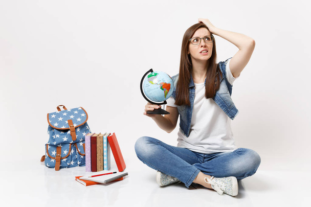 Young concerned woman student in glasses holding world globe clinging to head looking up sitting near backpack, school books isolated on white background. Education in high school university college - Foto, Imagem