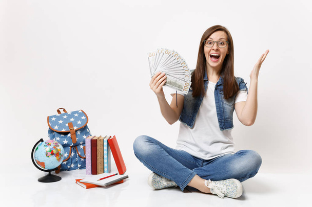 Молода збуджена студентка тримає пакет багато доларів, готівкові гроші роздають руки, сидячи поблизу рюкзака, шкільні книги ізольовані на білому тлі. Освіта в середній школі університетський коледж
 - Фото, зображення